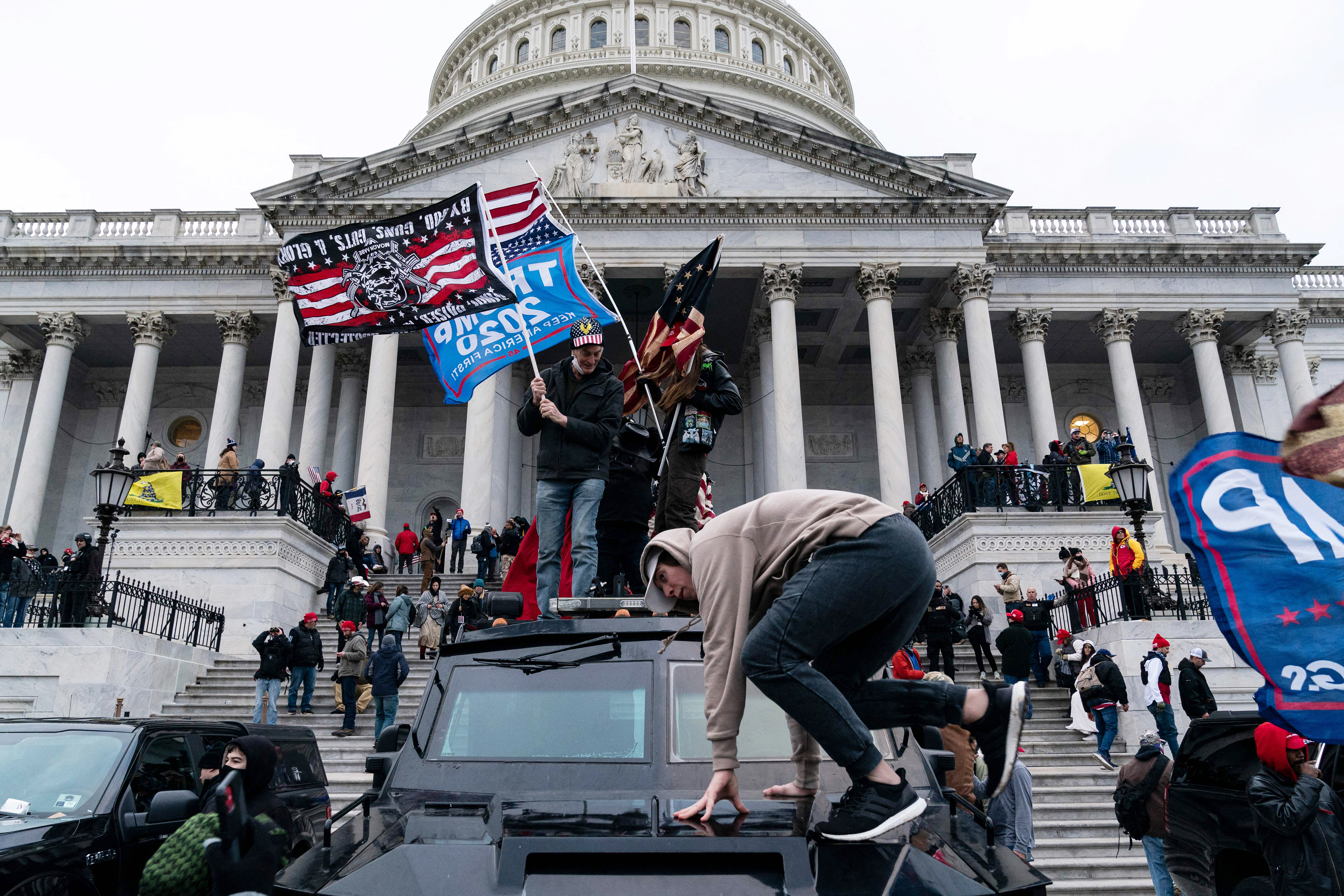 People waving flags while climbing on top of steps, military vehicles, and railings outside the Capitol building.